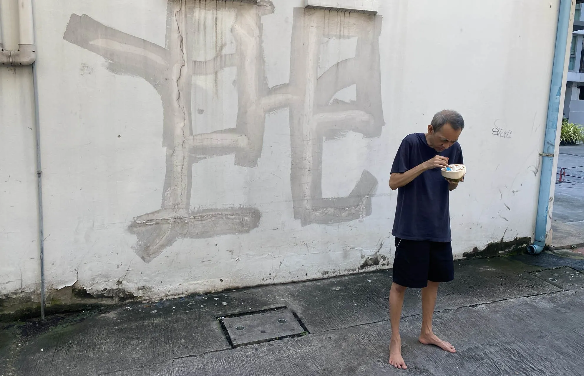 Man in a dark T-shirt eating from a coconut shell, standing barefoot in front of a white wall with faded lettering