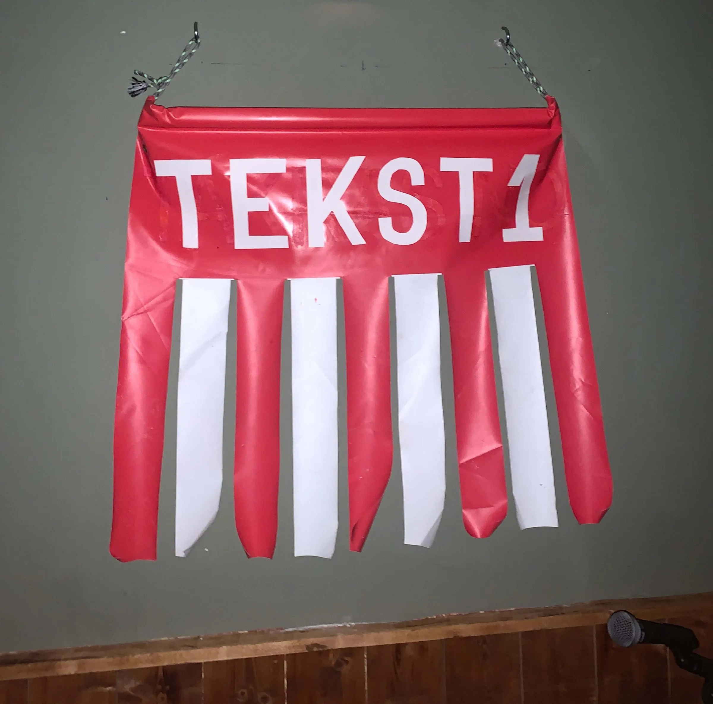Red banner reading TEKST1 with white cut strips, hanging on a wall above a stage