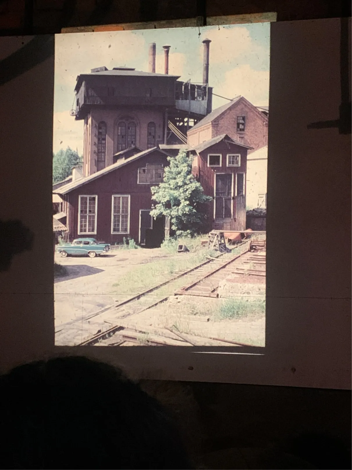 Projected slide of a run-down red brick and wooden industrial building with railway tracks and a vintage car