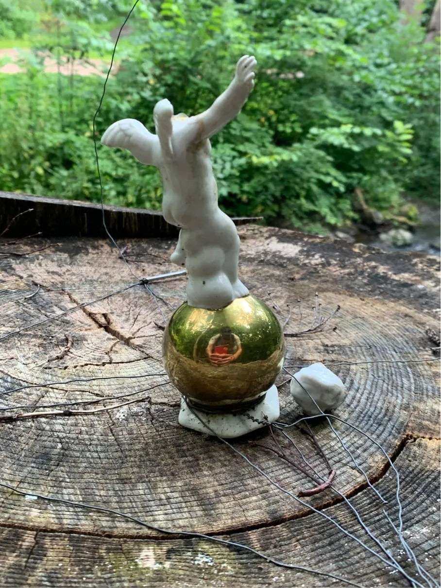 Small white porcelain figure balancing on a gold ball on a tree stump, surrounded by wire
