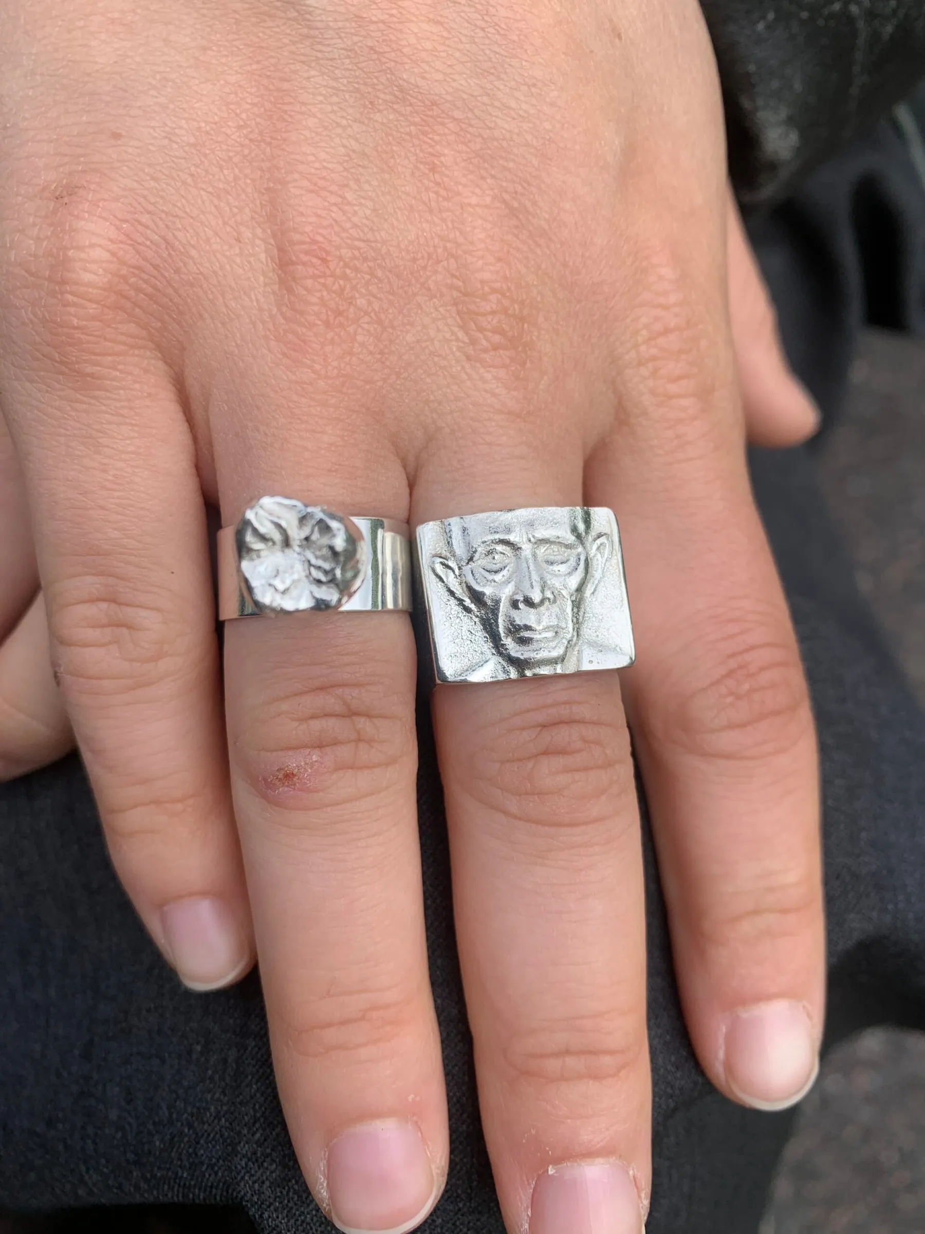 Hand wearing two silver rings, one with a carved face and one with a crumpled relief