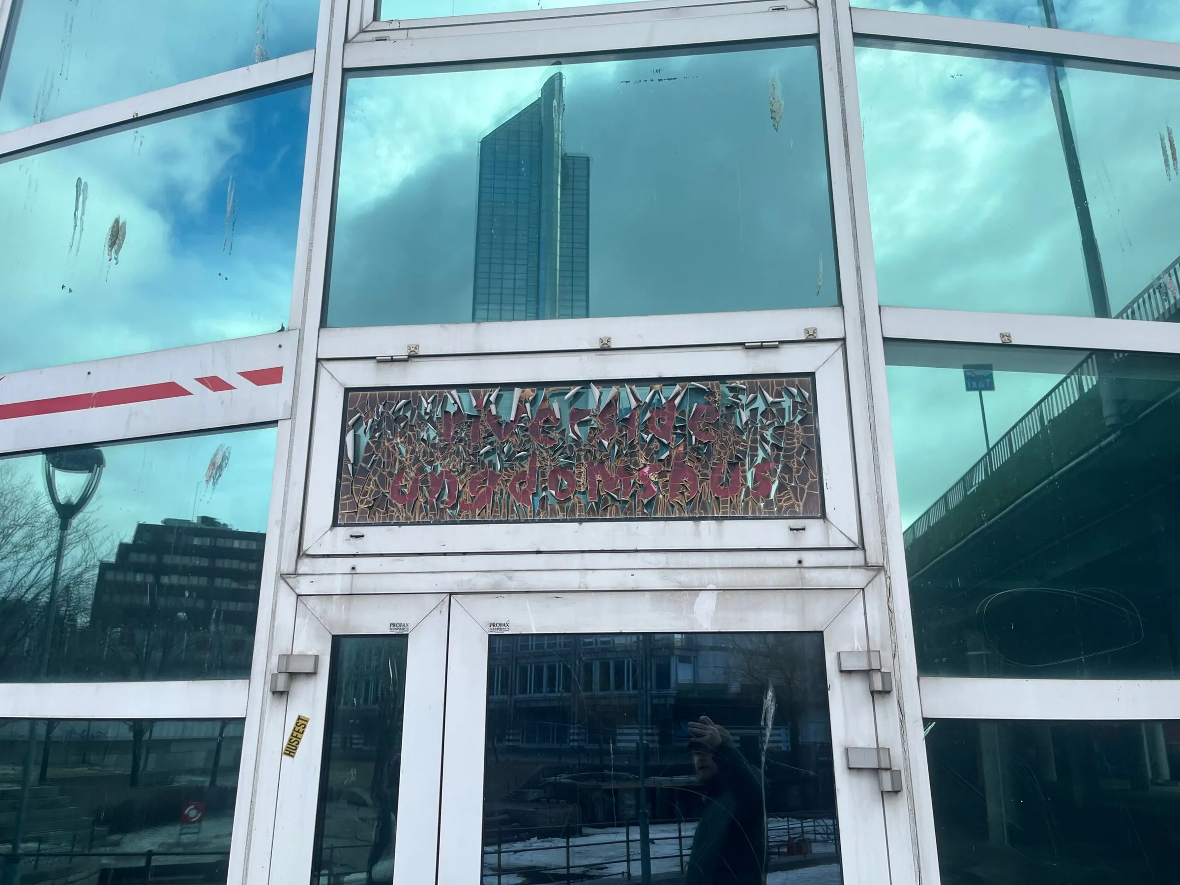 Glass facade and doorway with a cracked mosaic sign reading Riverside ungdomshus and a tower reflected above
