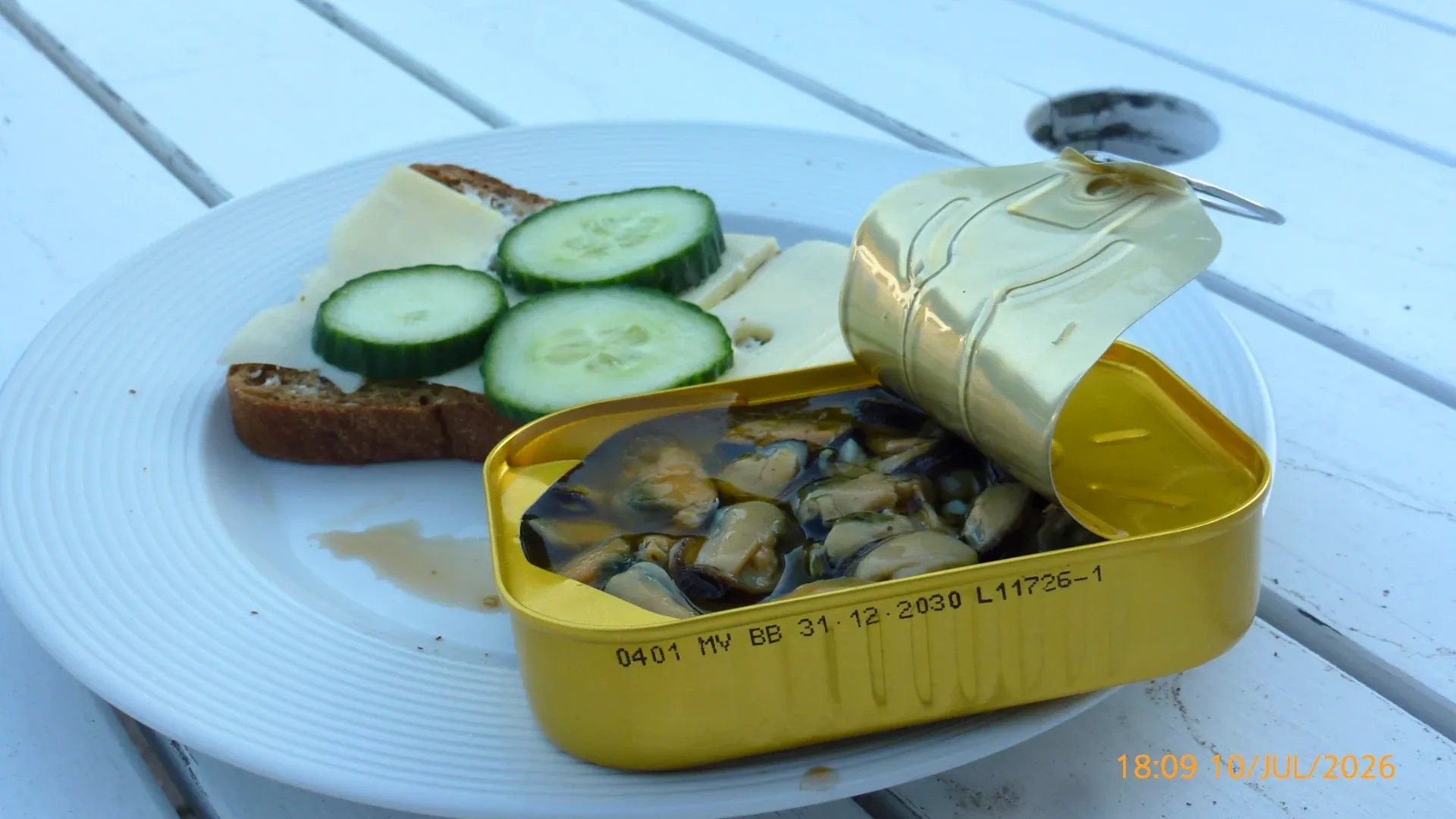 Open tin of mussels beside a slice of bread with cheese and cucumber on a white plate