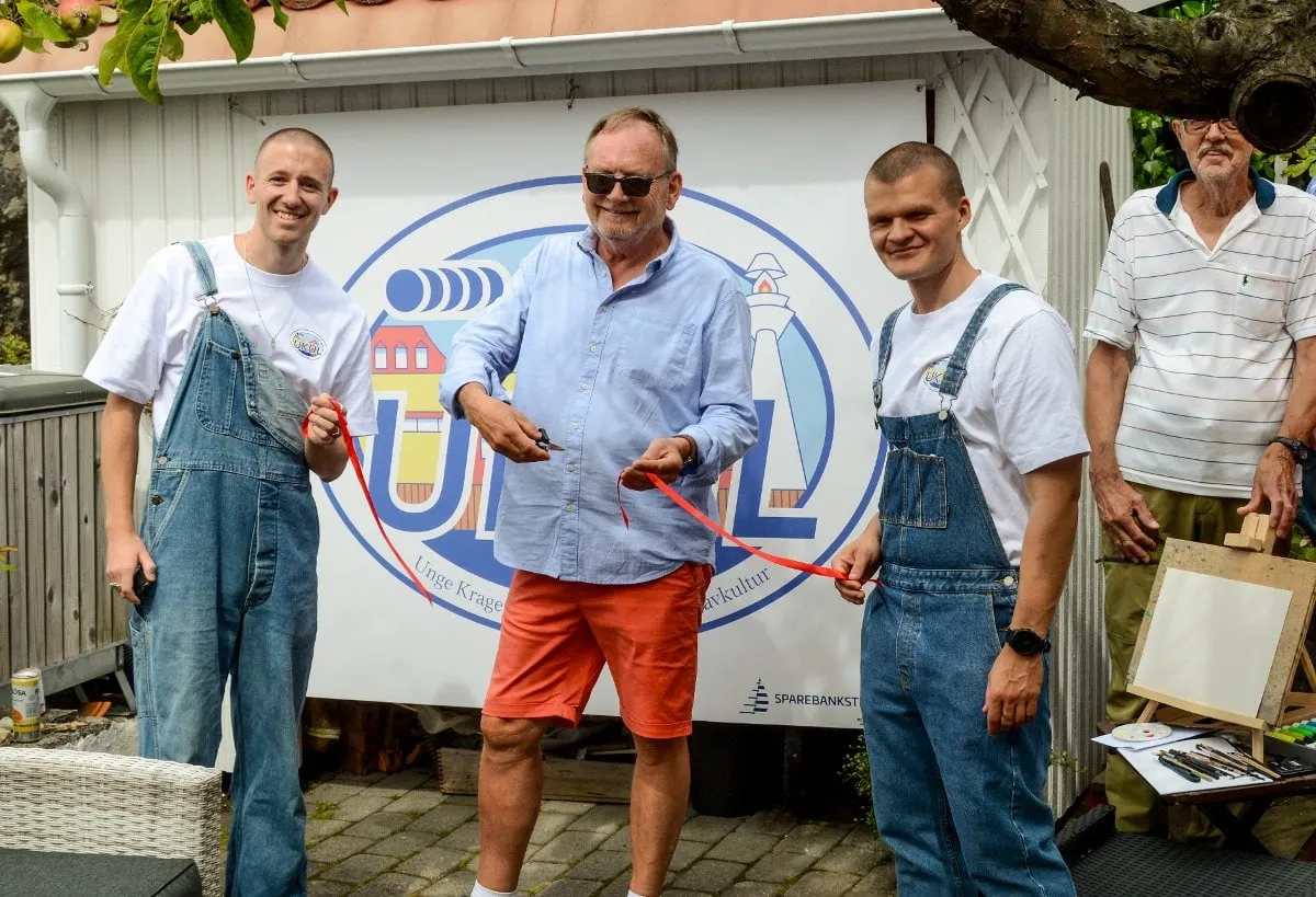Man in blue shirt cutting a red ribbon in front of the UKUL banner, flanked by two men in overalls and an older man
